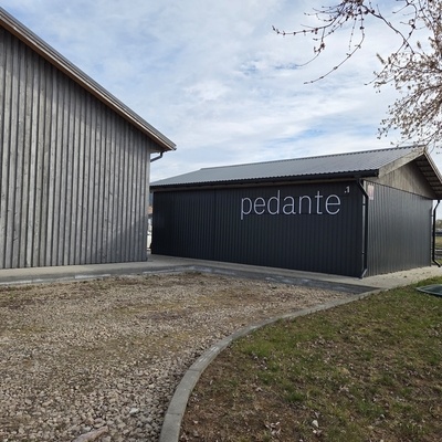 Vue extérieure de l’atelier Pedante en Lettonie, avec le logo Pedante sur un bâtiment de production à bardage métallique sombre.
